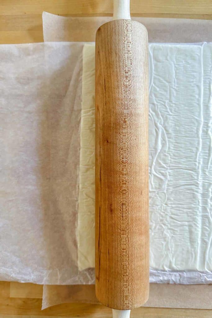 A wooden rolling pin on a wooden table rolling puff pastry.
