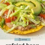 A bean tostada topped with refried beans, shredded lettuce, tomato, cheese, and sliced avocado on a white plate. Text reads "refried bean tostadas.