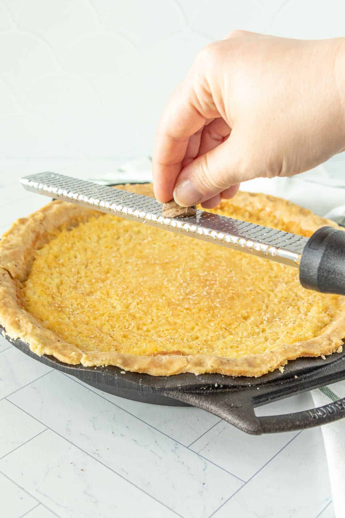 A person grating nutmeg over a pie in a cast iron skillet.