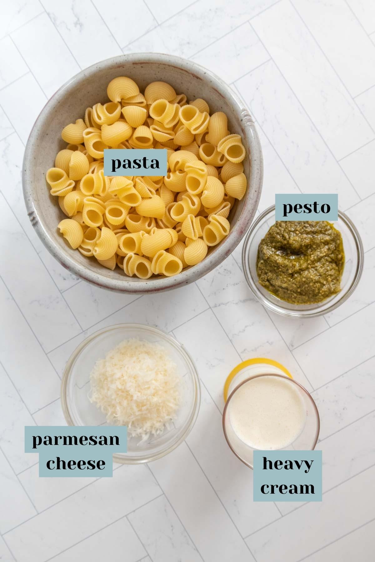 Ingredients for creamy pesto pasta on a tile surface with labels.