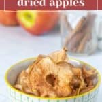 Bowl of dried apples and cinnamon on a white counter, with fresh apples and cinnamon sticks blurred in the background.
