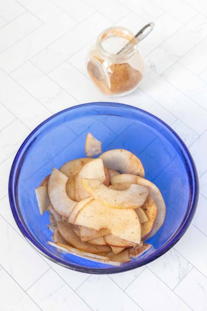 Apple slices in a blue bowl next to a bowl of cinnamon.