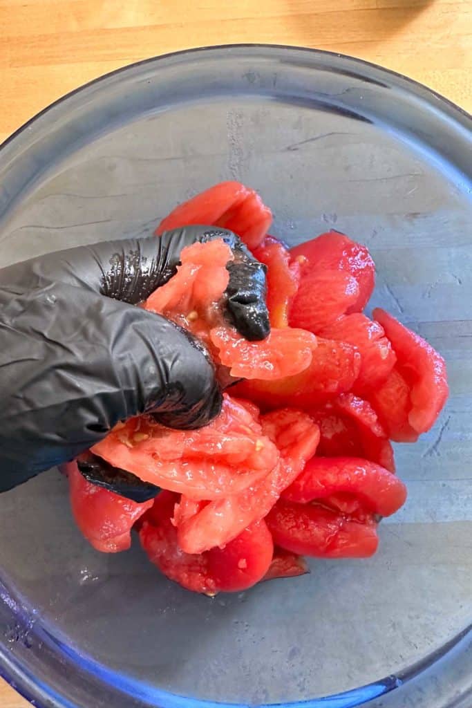 A person in black gloves crushing tomatoes in a bowl.