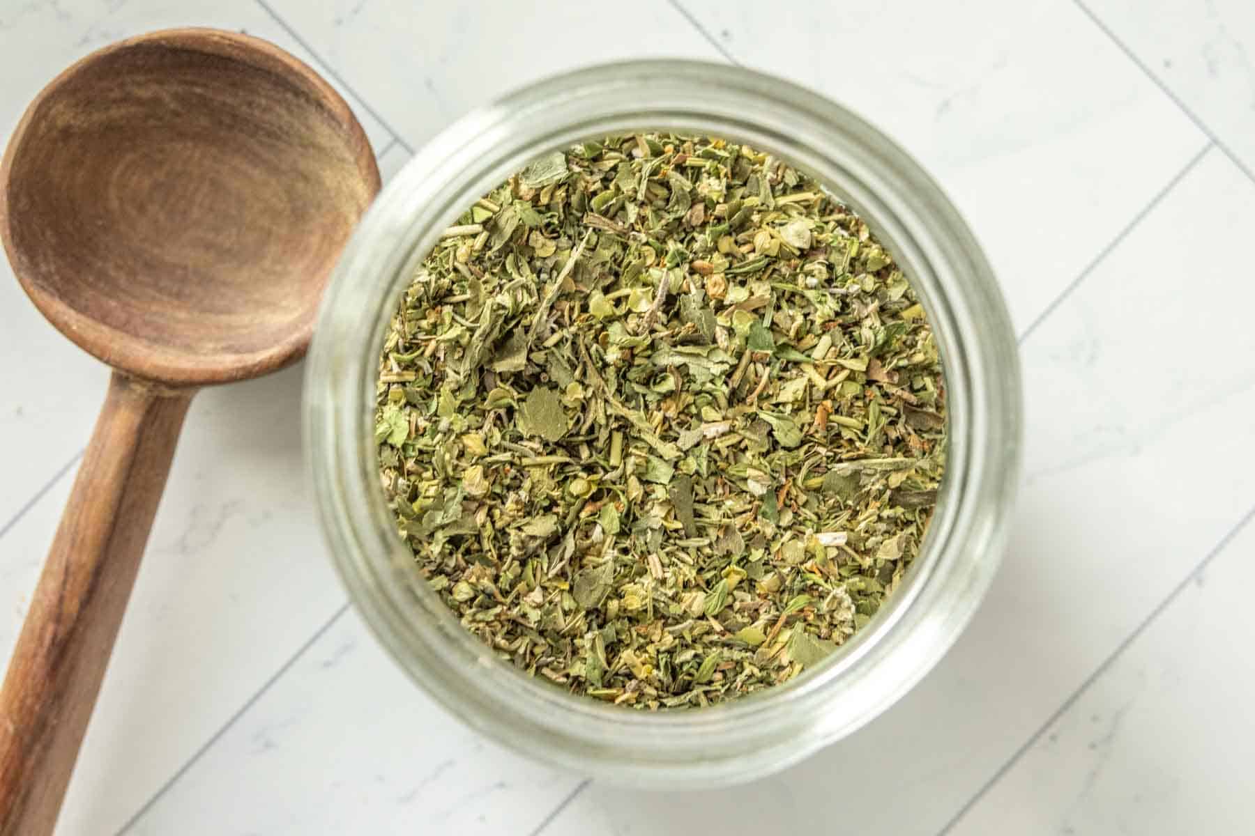 A jar of dried herbs next to a wooden spoon.