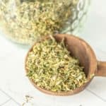 A wooden spoon full of dried herbs in front of a glass jar.