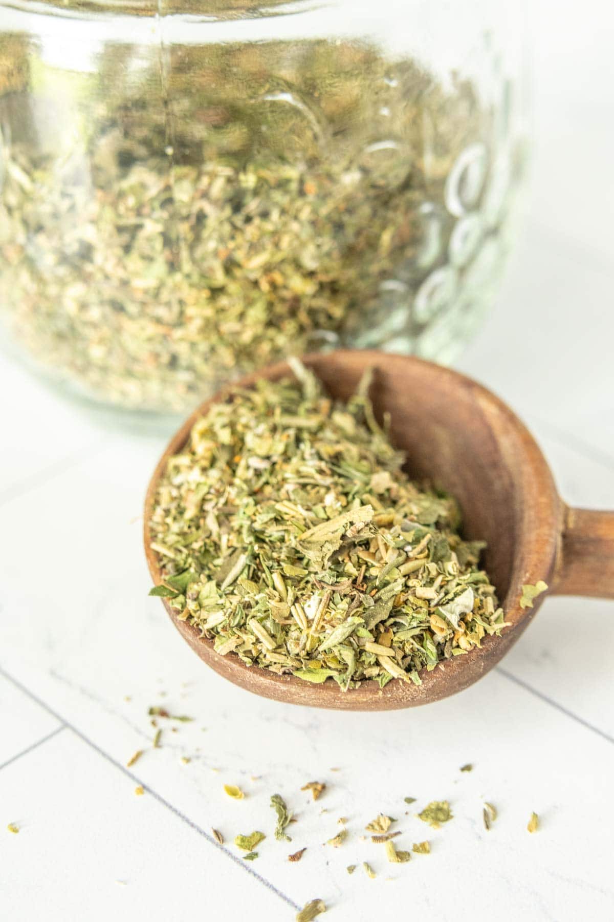 A wooden spoon full of dried herbs in front of a glass jar.