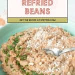 A bowl of creamy refried beans garnished with chopped herbs and grated cheese, with a spoon resting in the refried beans. Text overlay reads "Refried Beans – Get the recipe at stetted.com.