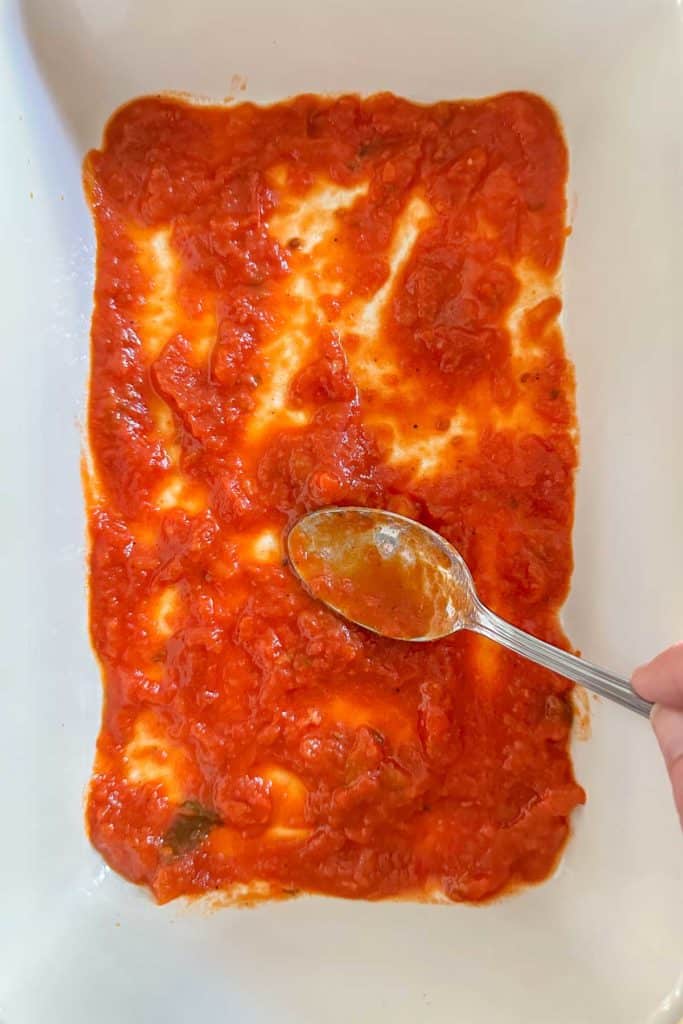 Spreading pasta sauce on the bottom of a casserole dish.