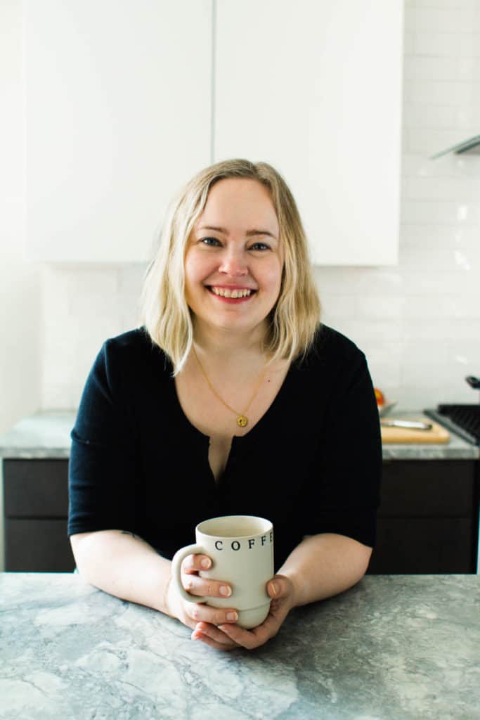 White woman in a black shirt leaning on countertop and holding a coffee mug.