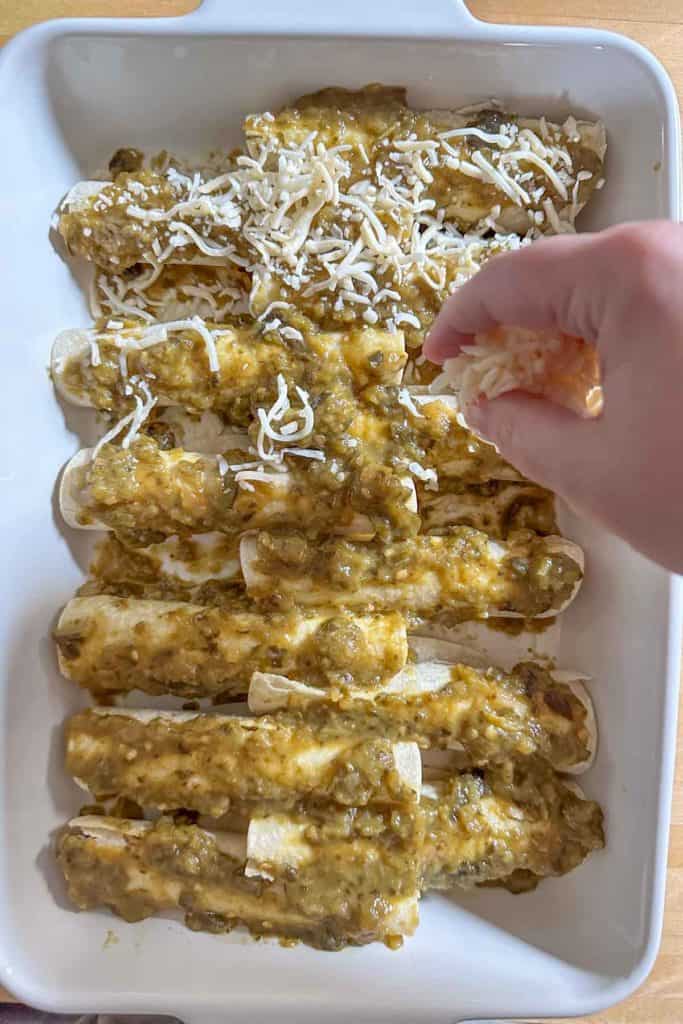 A person adding cheese to enchiladas into a baking dish.