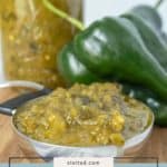A metal spoon filled with green chile sauce sits in front of fresh green chiles and a jar of homemade green chile sauce on a rustic wooden surface.