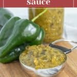 A metal spoon filled with green chile sauce rests on a wooden board beside a fresh green chile pepper and a jar of flavorful green chile sauce.