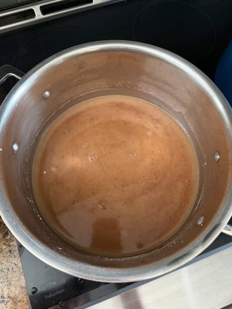 A stainless steel pot filled with a light brown apple pie filling mixture is heating on a stovetop.