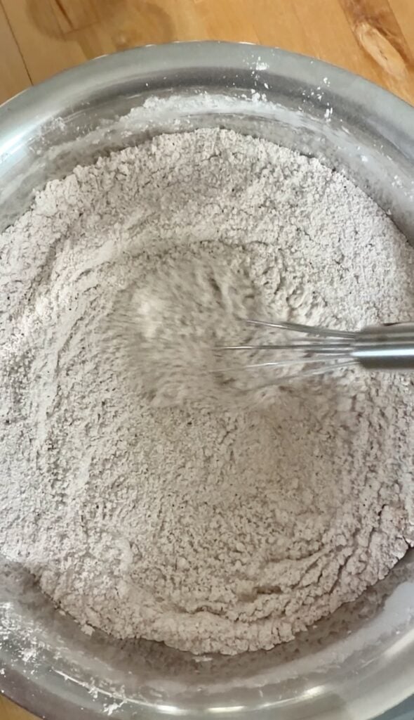 A metal whisk mixing dry ingredients, possibly flour and cocoa powder, in a metal bowl—preparing the base for a delicious dessert or perhaps the crust to pair perfectly with apple pie filling.