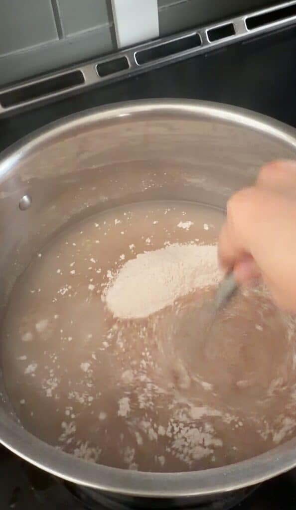 A hand stirs white powder into a pot of water on a stove, causing the powder to dissolve and create a cloudy mixture—the first step in preparing homemade apple pie filling.