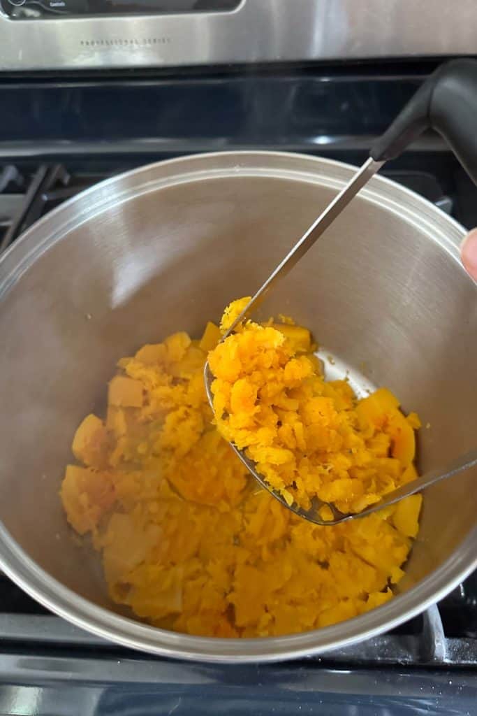 A person mashing squash in a pan on the stove.