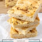 Three slices of fluffy no-knead focaccia are stacked on parchment paper, with more pieces of golden no-knead focaccia in the background on a wooden board.