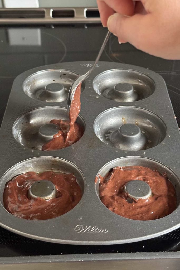 A person pouring chocolate batter into a donut tin.