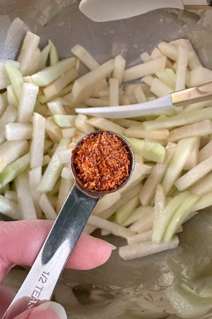 A person using a spoon to add seasoning to a bowl of jicama.