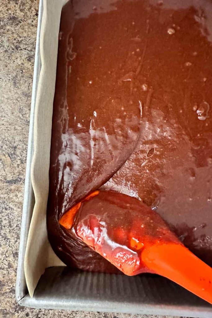 An orange spatula is being used to pour chocolate into a pan.