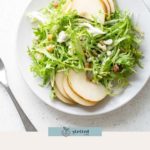A white plate with frisée salad topped with crumbled cheese, walnuts, and thin slices of pear sits on a white surface.