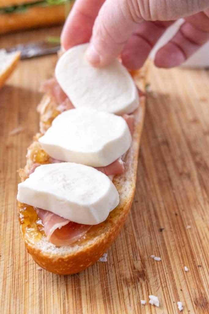 A person putting cheese on a bread with prosciutto.