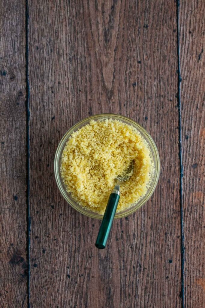 A glass bowl filled with yellow breadcrumbs and a green-handled fork, placed on a wooden surface—perfect ingredients for preparing a classic asparagus casserole.