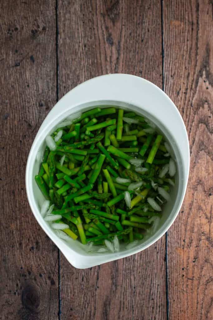 A bowl of chopped asparagus in water on a wooden surface.