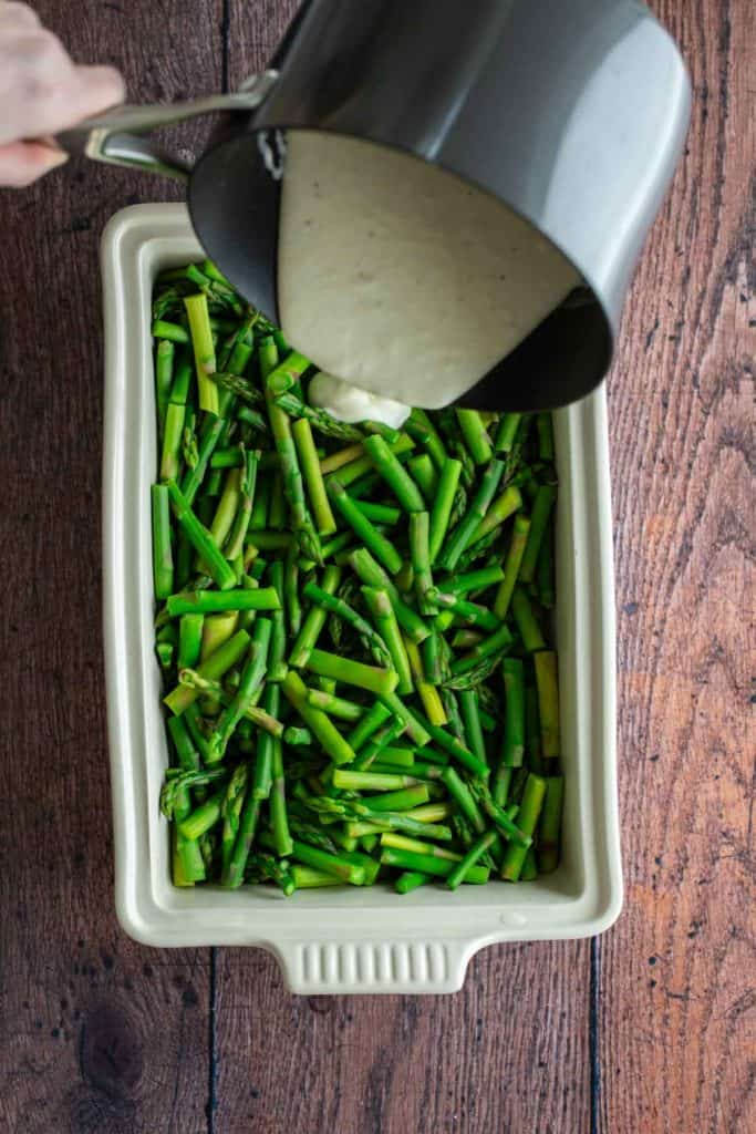 Pouring sauce over chopped asparagus in a baking dish.