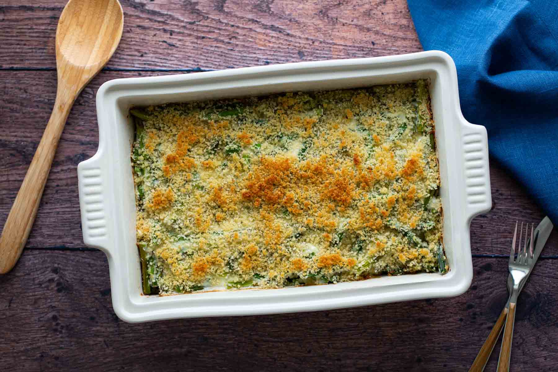 A freshly baked asparagus casserole in a white dish, with a golden breadcrumb topping, accompanied by a wooden spoon and a fork on a wooden table.