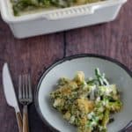 A serving of asparagus casserole on a plate with a fork and knife, beside a casserole dish.