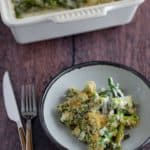 A serving of asparagus casserole on a plate with a fork and knife, beside a casserole dish.