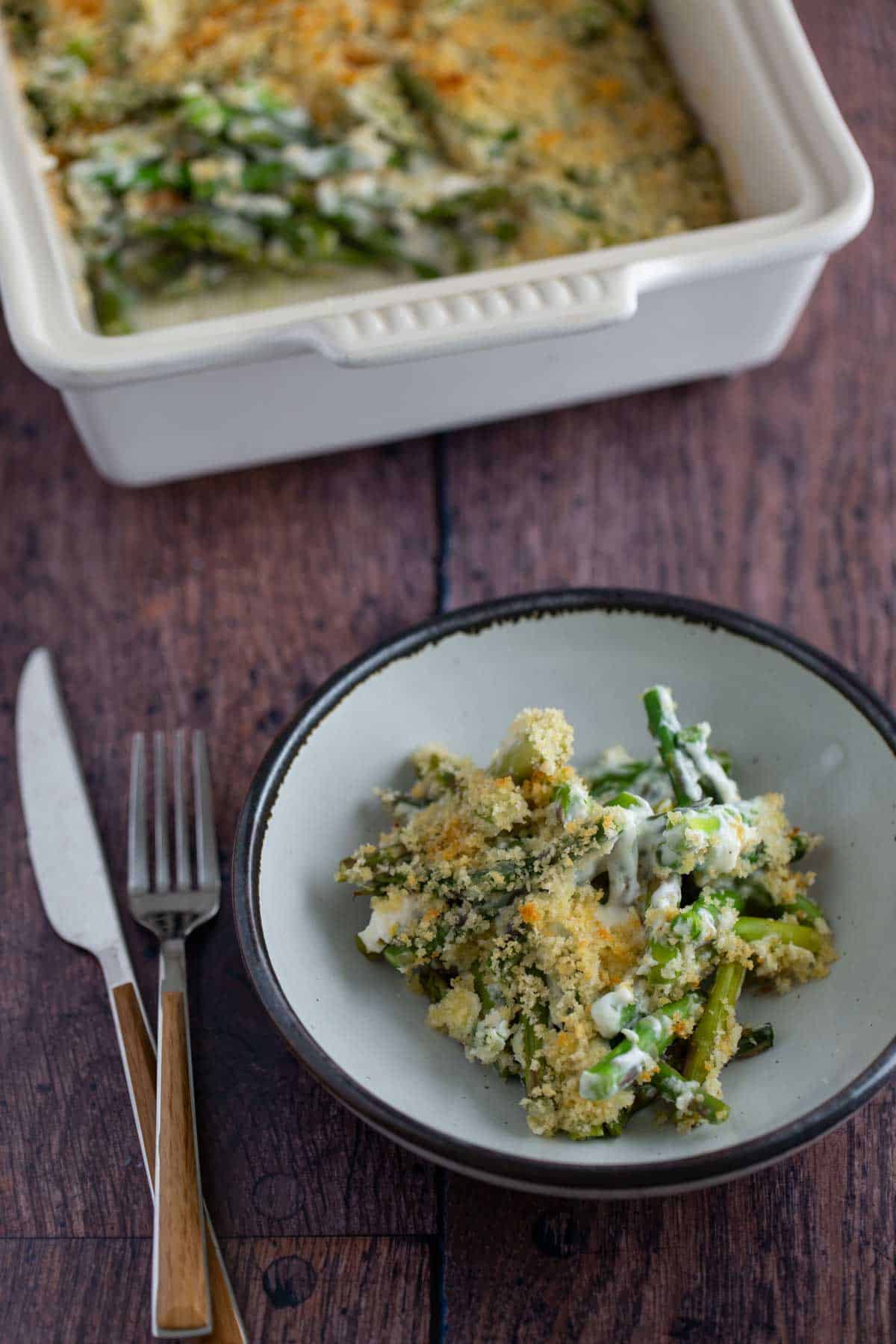 A serving of asparagus casserole on a plate with a fork and knife, beside a casserole dish.