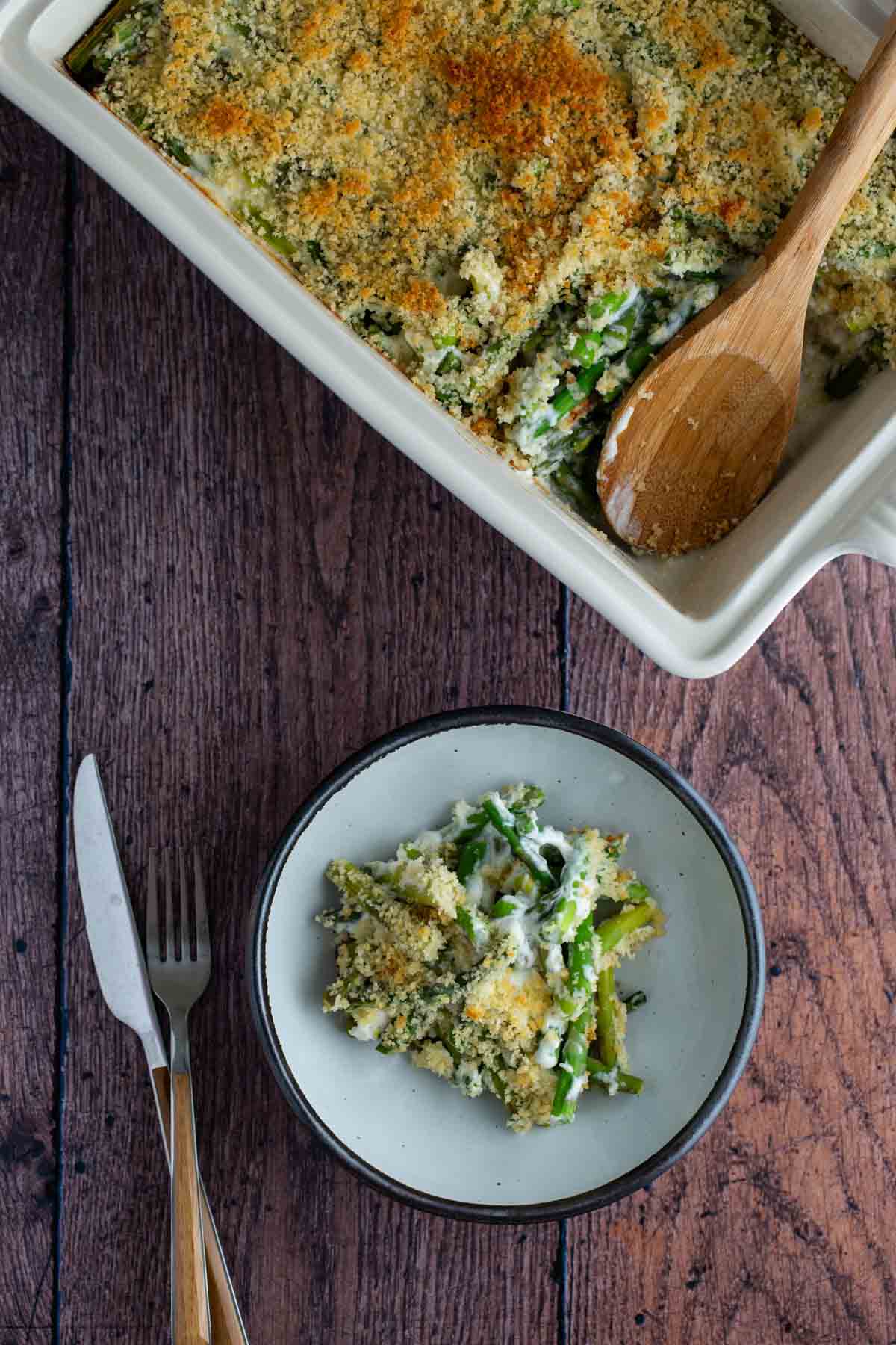 A serving of asparagus casserole topped with breadcrumbs in a ceramic dish with a wooden spoon, alongside a plated portion with a fork.