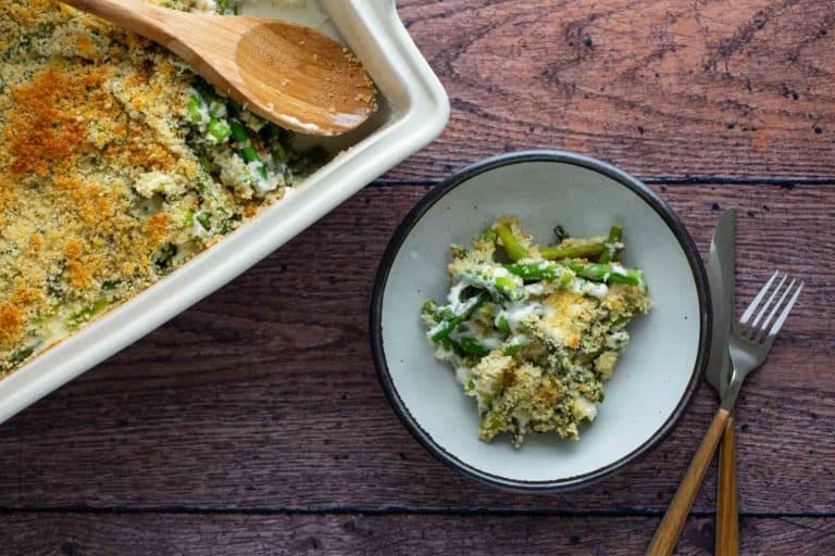A serving of asparagus casserole on a plate next to the casserole dish on a wooden table.