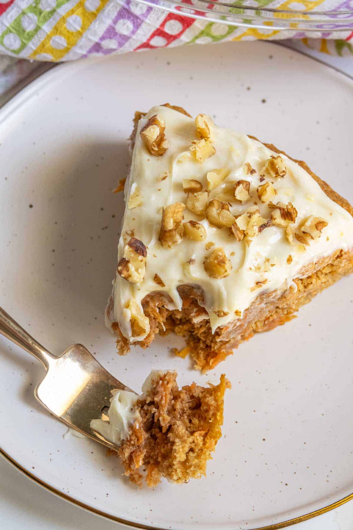 A slice of carrot cake with cream cheese frosting and chopped nuts on a plate with a fork.