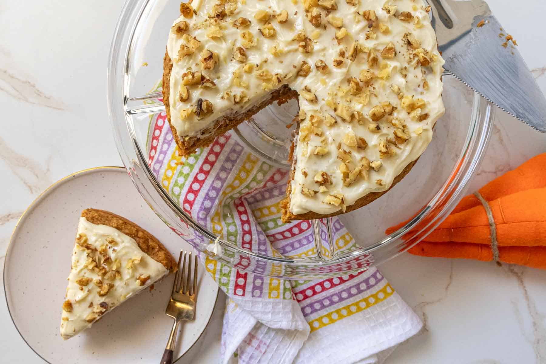 A carrot cake with cream cheese frosting and chopped nuts, with one slice served on a plate.