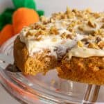 A carrot cake with cream cheese frosting and chopped nuts on a glass stand, with a few whole carrots in the background.