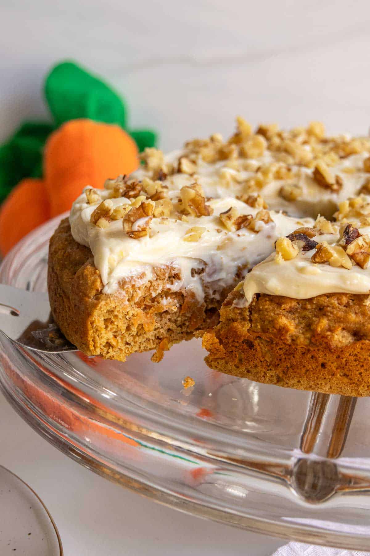 A carrot cake with cream cheese frosting and chopped nuts on a glass stand, with a few whole carrots in the background.