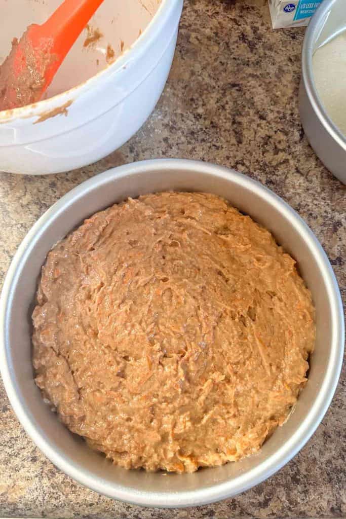 Unbaked cake batter in a round cake pan next to a mixing bowl and spatula.