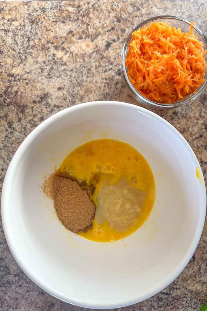 Ingredients for a recipe, including beaten eggs, brown sugar, and shredded carrots, arranged on a kitchen countertop.