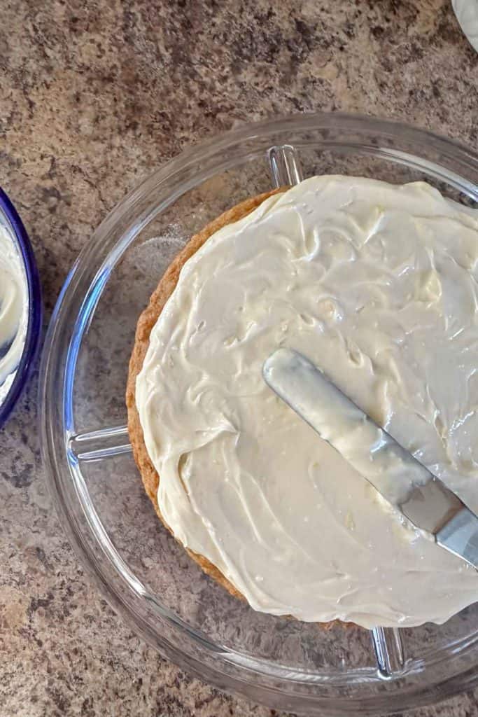 A cake with white frosting being smoothed with a spatula in a glass dish.