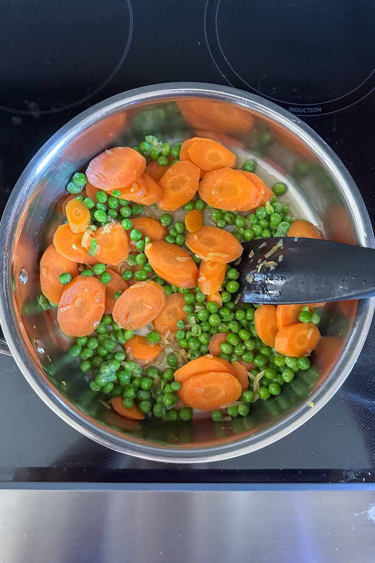 Sliced carrots and peas cooking in a stainless steel pot on an induction stove.