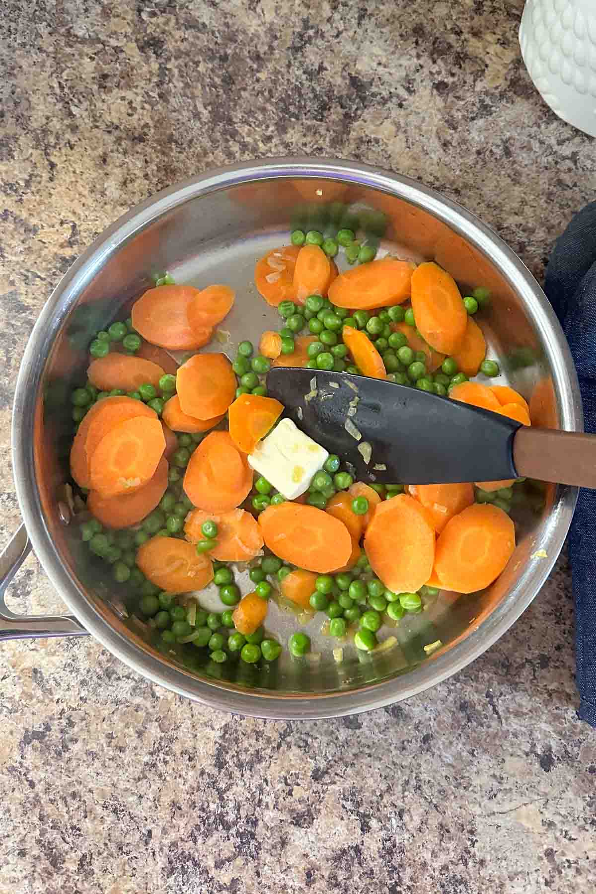 Sliced carrots and peas in a stainless steel pan with a spatula.