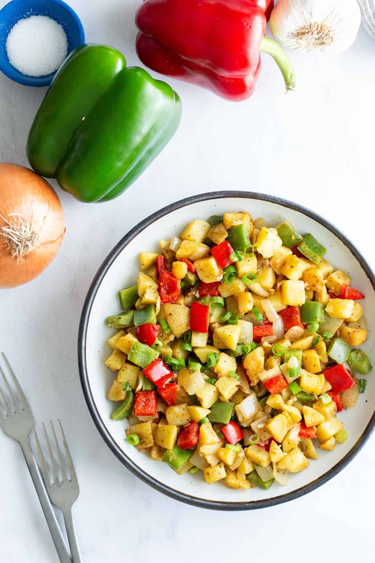 A plate of diced, sautéed potatoes with red and green bell peppers, accompanied by whole onions, garlic, and a bowl of salt on a white surface.