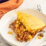 A serving of cornbread-topped beef and bean casserole on a white plate with a spoon, with the rest of the casserole visible in the background.