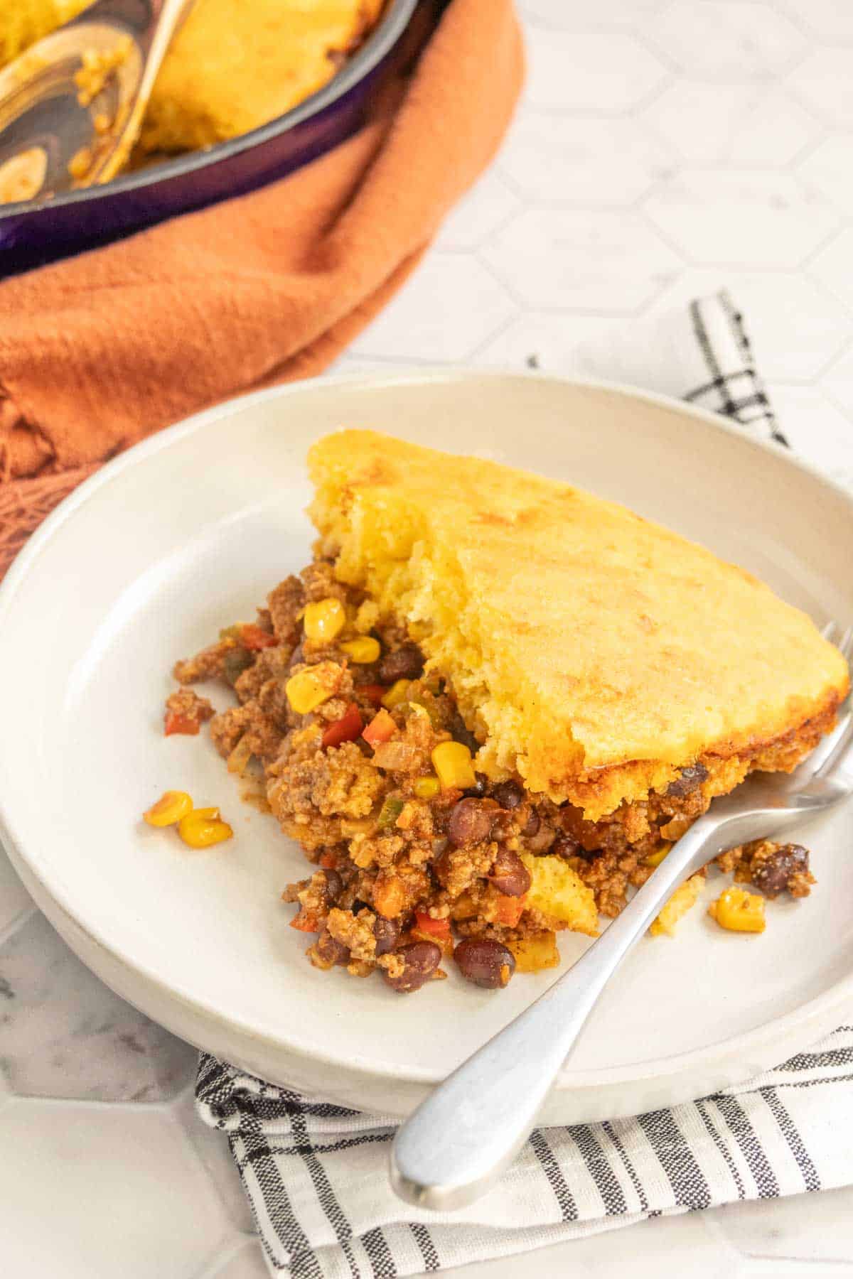 A serving of cornbread-topped beef and bean casserole on a white plate with a spoon, with the rest of the casserole visible in the background.