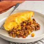 A slice of tamale pie casserole with ground meat, corn, beans, and a golden cornbread topping sits on a plate with a fork.