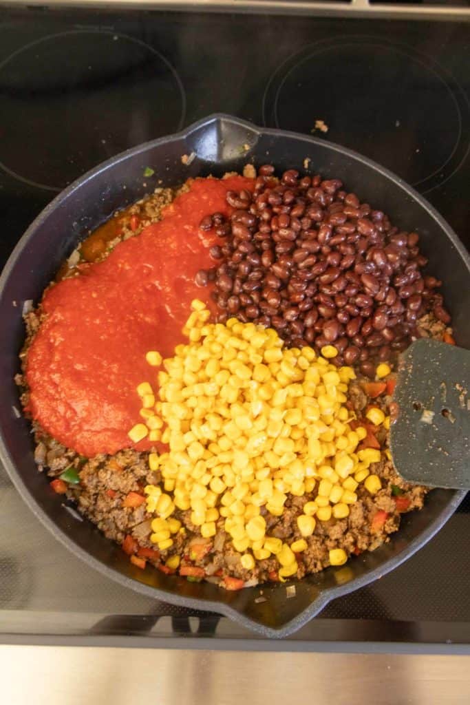 Cooking ingredients in a pan: ground meat, black beans, tomato sauce, and corn, mid-preparation.