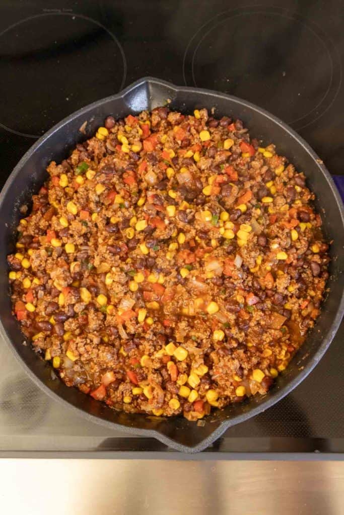 A skillet with cooked ground meat, beans, corn, and diced tomatoes on a stove top.
